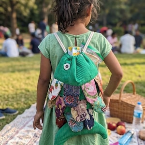 Op de afbeelding: Een groene rugzak in de vorm van een vis, met kleurrijke stoffen accenten, gedragen door een persoon in een groene jurk. De rugzak heeft witte bandjes en een enkel oog. De achtergrond toont een picknick.