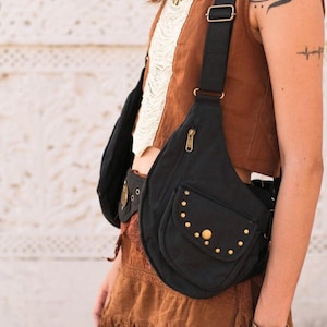 May include: A black canvas crossbody bag featuring a teardrop shape, an adjustable black strap, a vertical zippered compartment, and a front flap pocket adorned with small metallic studs and a central brass-toned button closure.