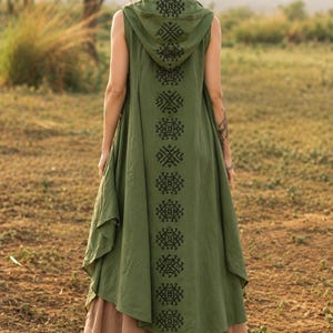 May include: An olive green sleeveless hooded vest featuring a long vertical row of black geometric embroidery running down the center back. The vest has an asymmetrical hemline and is layered over a tan, multi-layered flowing skirt.