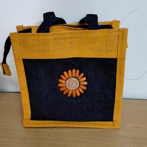 May include: A square jute tote bag with a black front panel and a sunflower design made of orange beads and a wooden button. The bag has a yellow border and black handles. The bag is approximately 8 inches wide and 8 inches tall.