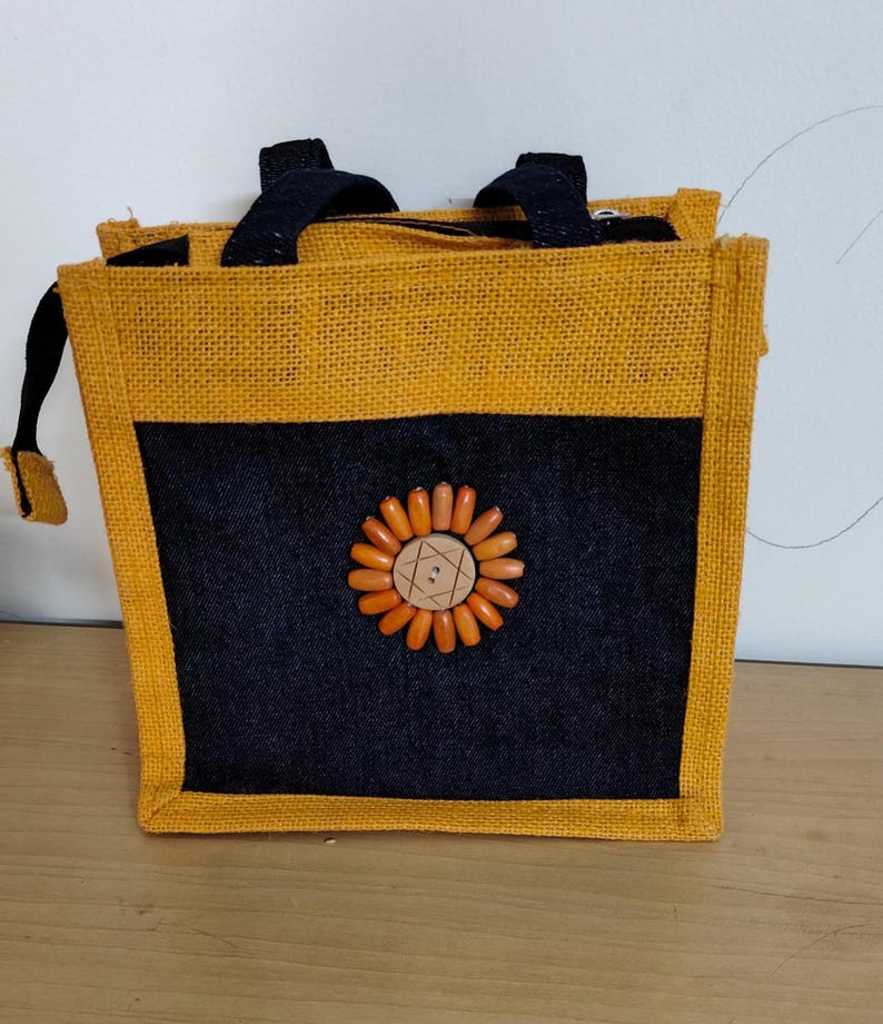 May include: A square jute tote bag with a black front panel and a sunflower design made of orange beads and a wooden button. The bag has a yellow border and black handles. The bag is approximately 8 inches wide and 8 inches tall.