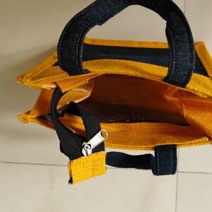 May include: A yellow tote bag with black accents. The bag has a rectangular shape with a zipper closure and two black handles. The interior of the bag is visible and appears to be a solid yellow color.