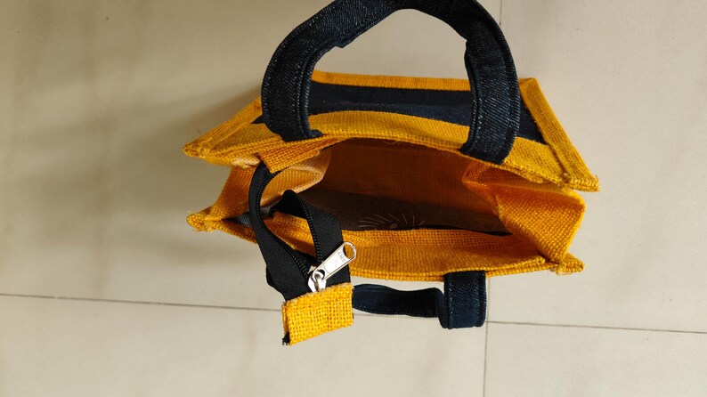May include: A yellow tote bag with black accents. The bag has a rectangular shape with a zipper closure and two black handles. The interior of the bag is visible and appears to be a solid yellow color.