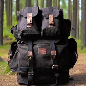 May include: A black canvas backpack with two top pockets and multiple compartments. The backpack features brown leather accents, adjustable straps, and metal buckles. The bag is designed for outdoor use and is set against a forest backdrop.