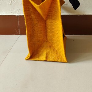 May include: A yellow burlap tote bag with black handles. The bag is rectangular with a structured shape and a visible woven texture. The handles are made of a dark, possibly navy, material.