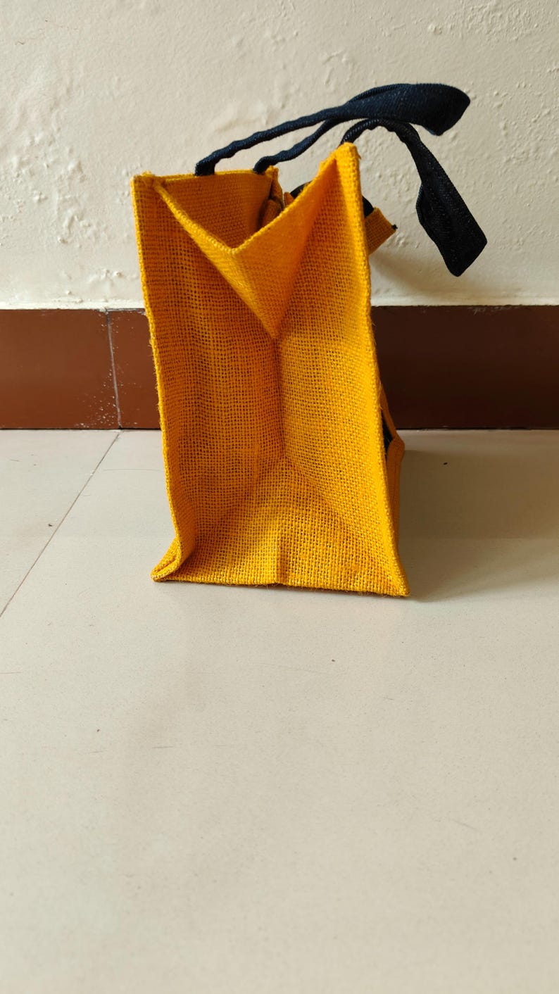 May include: A yellow burlap tote bag with black handles. The bag is rectangular with a structured shape and a visible woven texture. The handles are made of a dark, possibly navy, material.