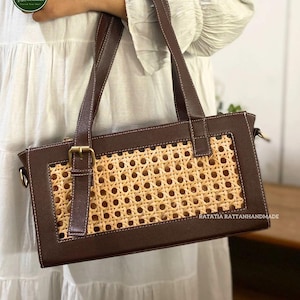 May include: A rectangular brown handbag with a woven rattan center panel. The bag has brown leather trim, handles, and a buckle closure. The bag is held by a person wearing a white dress. The bag has the text "RATATIA RATTANHANDMADE".