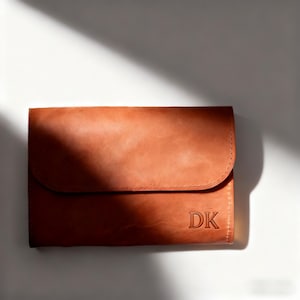 May include: A tan leather clutch bag with a curved flap closure. The initials "DK" are embossed on the lower right corner. The bag is set against a white background with a shadow.