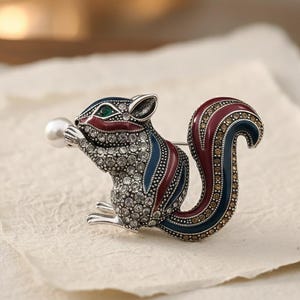 May include: Squirrel brooch with a pearl. The brooch is silver-toned with a detailed design. The squirrel has a red, blue, and brown tail. The body is covered in small, clear crystals. The squirrel is holding a white pearl.