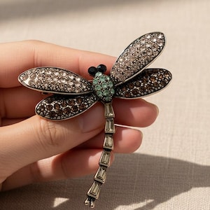 May include: A dragonfly brooch with crystal accents. The wings are adorned with clear and brown crystals, while the body features a green and black gemstone design. The brooch is made of metal and is a decorative accessory.