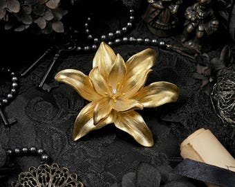 Peut inclure: Broche dorée en forme de lys avec une pierre précieuse centrale. L'image présente des clés anciennes, des perles et des éléments décoratifs sur fond noir. Un bijou élégant et raffiné.