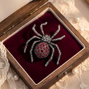 May include: A detailed spider brooch with a red and silver design, set in a wooden box with a red velvet interior. The spider's body is a cluster of red stones, and its legs are silver. The box is open, revealing the brooch.