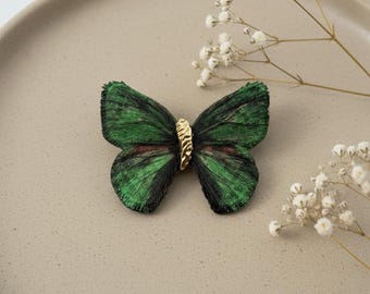 May include: A green and black butterfly brooch with a gold-colored center. The butterfly has detailed wing patterns and is set against a neutral background. Decorative floral accents are in the background.