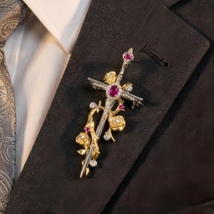 Könnte beinhalten: Aufwendige Brosche in Form eines Schwertes und Kreuzes, verziert mit einem großen zentralen Rubin und kleineren Rubinen. Die Brosche hat goldene und silberne Akzente, diamantähnliche Verzierungen und Rosenmotive. Sie ist an einem schwarzen Blazer befestigt.