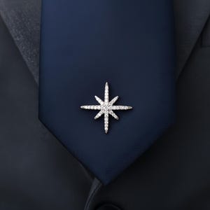 May include: A silver starburst brooch with clear crystals is pinned to a navy blue tie. The brooch has eight pointed rays and is set against a dark background, suggesting formal wear or an accessory.