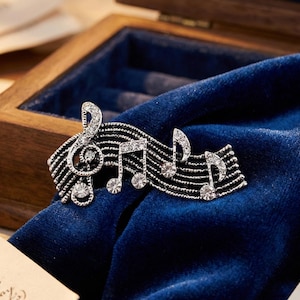 Puede incluir: Broche plateado con notas musicales y una clave de sol, adornado con cristales transparentes. El broche se presenta sobre un fondo de terciopelo azul oscuro, sugiriendo un tema de joyería o accesorios. El diseño incorpora símbolos musicales.