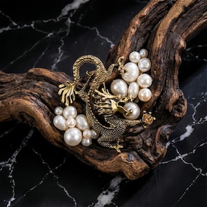 May include: A gold-toned dragon brooch with intricate detailing, adorned with clusters of white pearls and a single amber-colored gem. The brooch is set against a textured, dark wooden surface with a black marble background.