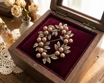 May include: A vintage-style brooch with a wreath design, featuring faux pearls and leaf accents. The brooch is displayed inside a wooden jewelry box with a burgundy velvet lining. The box is open, revealing the brooch's intricate details.