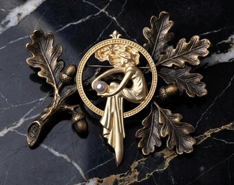 May include: A gold-coloured brooch depicting a woman holding a pearl within a circular frame. The design incorporates oak leaves, acorns, and a branch, set against a black marble background. The brooch has a vintage aesthetic.