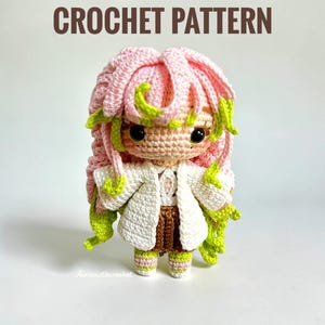 Pode incluir: Boneca de crochê com cabelo rosa e detalhes verdes, vestida com um casaco branco e uma saia marrom. A boneca tem olhos grandes e pretos e está em pé. O texto "CROCHET PATTERN" está acima da boneca.
