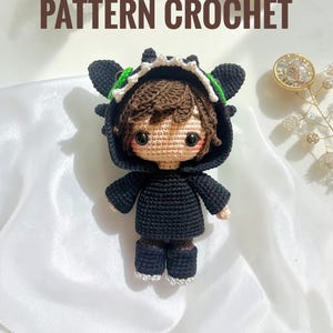 Puede incluir: Muñeca de ganchillo hecha a mano con un disfraz de dragón negro con detalles verdes. La muñeca tiene cabello castaño y mejillas rosadas. El texto "PATTERN CROCHET" se muestra encima de la muñeca. Sobre una superficie de satén blanco.