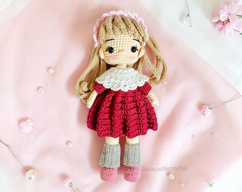 May include: A handmade crochet doll with long blonde hair, wearing a red dress, gray socks, and pink shoes. The doll has a white collar and a pink headband. The text "PATTERN CROCHET" is displayed above the doll.