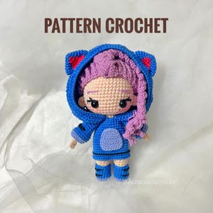 Peut inclure: Poupée au crochet faite à la main, vêtue d'une veste à capuche bleue avec des oreilles de chat et des bottes assorties. La poupée a des cheveux violets et un teint clair. Le texte "PATTERN CROCHET" est affiché au-dessus de la poupée.