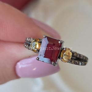 Könnte beinhalten: Ein Ring mit einem zentralen, smaragdgeschliffenen, tiefroten Edelstein, flankiert von zwei ovalen gelben Steinen. Die Ringschiene besteht aus dunklem Metall mit kleinen Pavé-Steinen an den Seiten. Der Text ElegantJewelry ist zu sehen.
