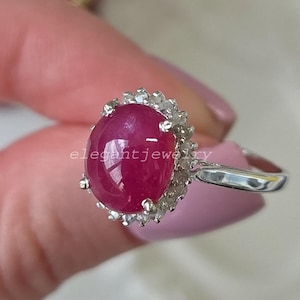 Könnte beinhalten: Ein Silberring mit einem großen, ovalen, im Cabochon-Schliff gehaltenen, tiefrosa Rubin in der Mitte. Der Rubin ist von einem Kranz aus kleinen, klaren, rund geschliffenen weißen Steinen in Silberfassung umgeben. Der Text "elegantjewelry" ist sichtbar.