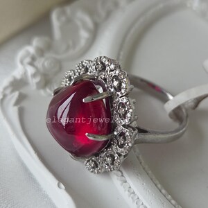 Könnte beinhalten: Ein silberfarbener Ring mit einem großen, glatten, tiefroten Cabochon-Edelstein in einer Krappenfassung, umgeben von einem Kranz aus kleinen, klaren, funkelnden Steinen. Der Text "elegantjewel" ist auf dem roten Edelstein zu sehen.