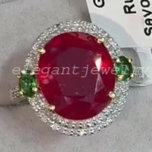 Könnte beinhalten: Ein Ring mit einem großen, oval geschliffenen Rubin, umgeben von einem Kranz aus kleineren Diamanten. Zwei grüne Edelsteine flankieren den Rubin. Der Ring ist in einem Goldband gefasst. Der Ring wird in einer Schmuckschatulle präsentiert.