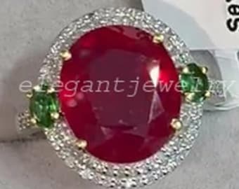 Puede incluir: Un anillo con un gran rubí de corte ovalado, rodeado por un halo de diamantes más pequeños. Dos gemas verdes flanquean el rubí. El anillo está engastado en una banda de oro. El anillo se exhibe en una caja de joyería.