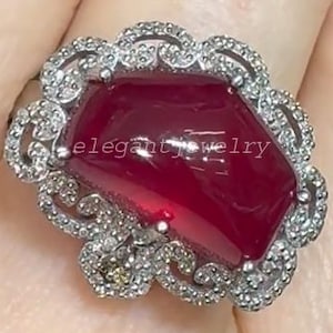 Könnte beinhalten: Ein silberner Ring mit einem großen, facettierten, tiefroten Edelstein. Der Edelstein ist von einem Kranz aus kleineren, klaren Steinen umgeben. Der Ring trägt den Text "elegant jewelry" auf dem Edelstein.