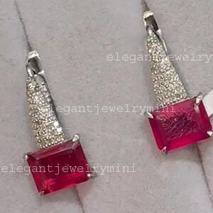 May include: A pair of silver earrings with a rectangular, vibrant red gemstone at the base. The upper portion of each earring is cone-shaped and encrusted with small, sparkling stones. The earrings are displayed on a light gray surface.