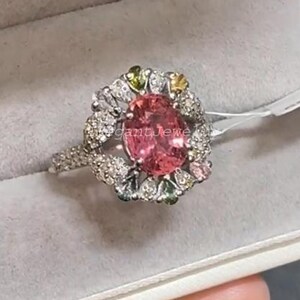 Könnte beinhalten: Ein Ring mit einem großen, oval geschliffenen rosa Edelstein in der Mitte, umgeben von kleineren weißen und farbigen Edelsteinen. Der Ring hat ein mit kleinen, klaren Steinen besetztes Band. Der Ring wird in einer grauen Schmuckschatulle präsentiert.