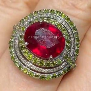 Könnte beinhalten: Nahaufnahme eines Rings mit einem großen, oval geschliffenen Rubin. Der Rubin ist von einem Kranz aus kleineren, runden grünen Edelsteinen umgeben und in einem silbernen Band gefasst. Der Text "elegant jewelry" ist auf dem Ring zu sehen.