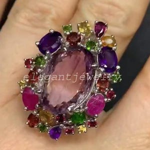 Könnte beinhalten: Ein auffälliger Ring mit einem großen, ovalen, rosa Edelstein in der Mitte, umgeben von einer Vielzahl kleinerer Edelsteine in Lila, Rot, Gelb und Grün. Der Ring ist in Silber oder Weißmetall gefasst. Der Text "elegant jewelry" ist sichtbar.