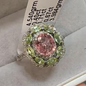 Op de afbeelding: Zilveren ring met een grote roze middensteen, omringd door kleinere groene en heldere stenen. De ring wordt gepresenteerd met een label met tekst. De ring zit in een doos.