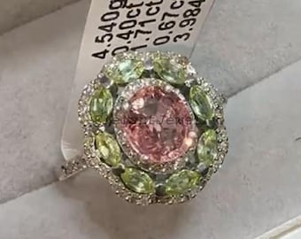 Puede incluir: Anillo plateado con una gran piedra rosa central, rodeada de piedras verdes y transparentes más pequeñas. El anillo se presenta con una etiqueta con texto. El anillo está en una caja.