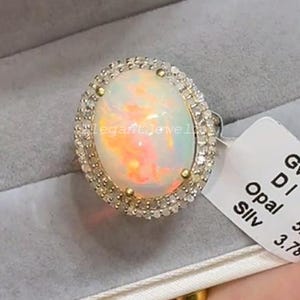 Könnte beinhalten: Ovaler Opalring mit einem Kranz aus kleinen Diamanten. Der Opal hat eine Mischung aus Farben, darunter Weiß, Orange und Rosa. Der Ring ist in einem Silberband gefasst. Der Ring befindet sich in einer grauen Schmuckschatulle.