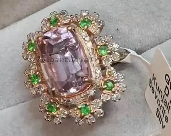 Puede incluir: Un anillo de oro rosa con una gran piedra preciosa de color púrpura claro en el centro. La piedra preciosa está rodeada por un halo de pequeñas piedras transparentes y gemas verdes. El anillo está sobre un fondo gris suave.