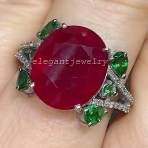 Könnte beinhalten: Ein silberner Ring mit einem großen, oval geschliffenen Rubin. Der Rubin ist von kleineren, birnenförmigen grünen Edelsteinen und kleinen weißen Edelsteinen umgeben. Der Text "elegant jewelry" ist auf dem Ring zu sehen.