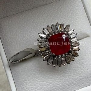 Puede incluir: Un anillo de tono plateado con una piedra roja central de corte cuadrado rodeada por un halo de piedras claras de corte baguette dispuestas en forma de sol. El anillo se presenta en un joyero blanco con el texto elegantjewelry visible.