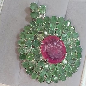 Könnte beinhalten: Ein Anhänger mit einem großen, ovalen, rubinroten Edelstein in der Mitte, umgeben von einem Kranz aus kleinen, grünen Smaragden. Der Anhänger ist in einem silberfarbenen Metall gefasst und wird in einer weißen Schmuckschatulle präsentiert.