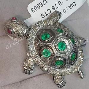 May include: A bejeweled turtle pendant with a silver-tone setting. The turtle's shell features emerald-green stones, and the head has a ruby eye. The pendant is set in a white box. The tag reads: AT: TS-476003, ST: 0.76 CTS, DIA: 0.5.