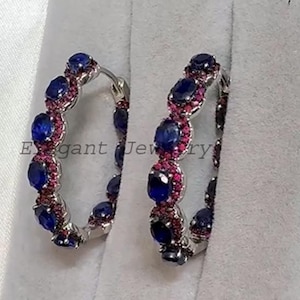 May include: A pair of silver hoop earrings, each featuring alternating oval-cut blue and red gemstones. The earrings are presented in a white jewellery box with a grey interior. The text "Elegant Jewelry" is visible.