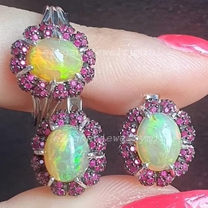 May include: A silver ring and earrings set featuring oval opals surrounded by small, round, pink gemstones. The opals have a mix of yellow and green hues. The earrings and ring have a floral design.