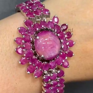 Puede incluir: Una pulsera plateada con una gema rosa grande y redonda, rodeada de gemas rosas ovaladas más pequeñas. La banda de la pulsera también está adornada con las mismas gemas rosas. La joya se muestra en una muñeca.