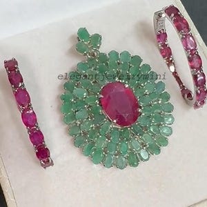 Op de afbeelding: Een sieradenset met een hanger en twee bijpassende oorbellen. De hanger heeft een groot ovaal robijn midden omgeven door smaragden. De oorbellen en hanger hebben de tekst "elegantjewelrymini".