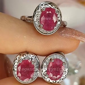 Könnte beinhalten: Ein silbernes Ring- und Ohrring-Set. Jedes Schmuckstück hat einen großen, ovalen, tiefroten Edelstein, der von kleinen, klaren Akzentsteinen umgeben ist. Der Ring und die Ohrringe haben ein dekoratives, durchbrochenes Design.